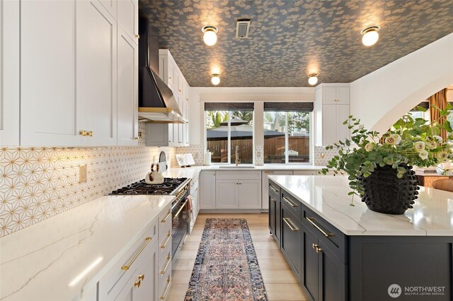 kitchen with wall chimney range hood, high end stainless steel range, white cabinets, a sink, and decorative backsplash