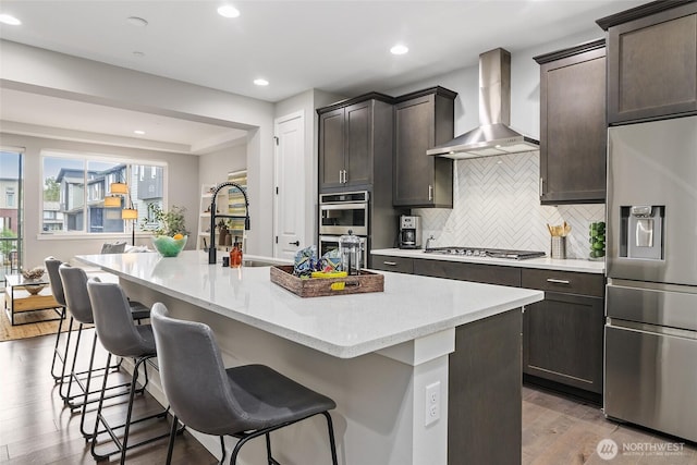 kitchen featuring stainless steel appliances, wall chimney range hood, a sink, wood finished floors, and a breakfast bar