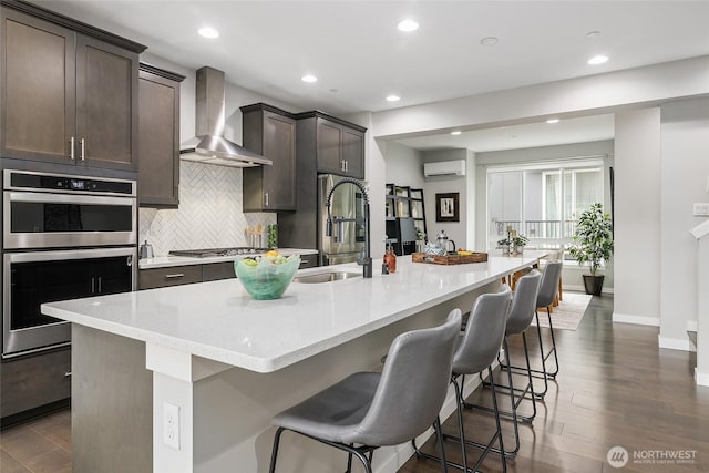 kitchen with wall chimney range hood, a wall mounted AC, stainless steel double oven, backsplash, and a sink