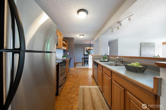 kitchen with appliances with stainless steel finishes, a textured ceiling, a sink, brown cabinetry, and a chandelier
