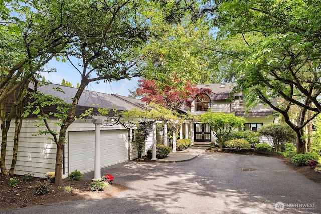 view of property hidden behind natural elements featuring asphalt driveway, an attached garage, and a shingled roof