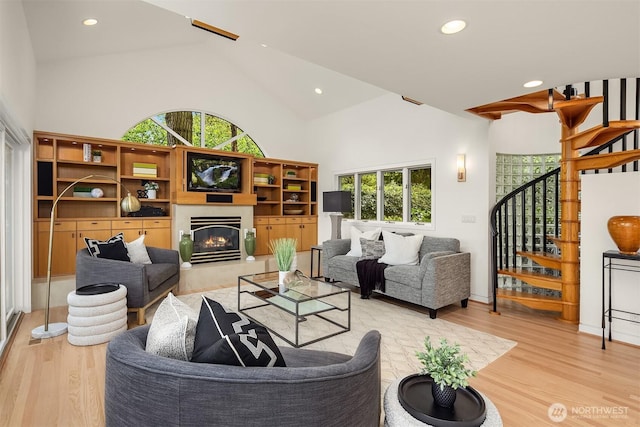 living area featuring stairway, light wood-style flooring, high vaulted ceiling, recessed lighting, and a glass covered fireplace