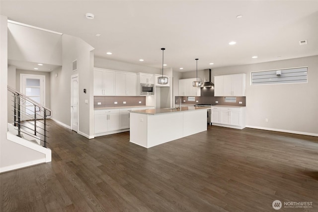 kitchen featuring appliances with stainless steel finishes, wall chimney exhaust hood, tasteful backsplash, baseboards, and a center island with sink
