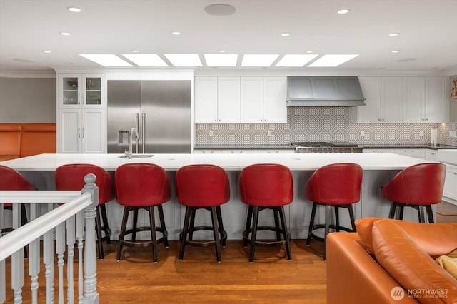 kitchen featuring custom exhaust hood, high end fridge, a sink, tasteful backsplash, and a large island with sink