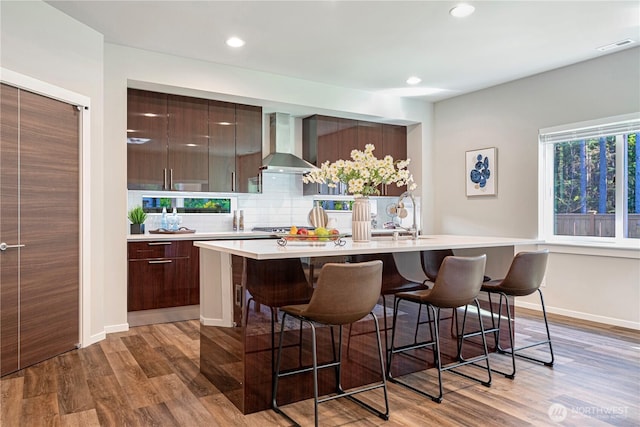 kitchen featuring wall chimney exhaust hood, modern cabinets, light countertops, a breakfast bar area, and recessed lighting