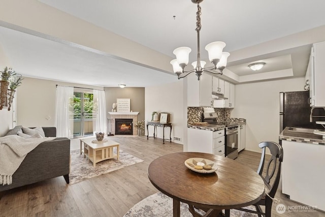 dining space featuring light wood-style floors, a chandelier, a warm lit fireplace, baseboards, and a tray ceiling