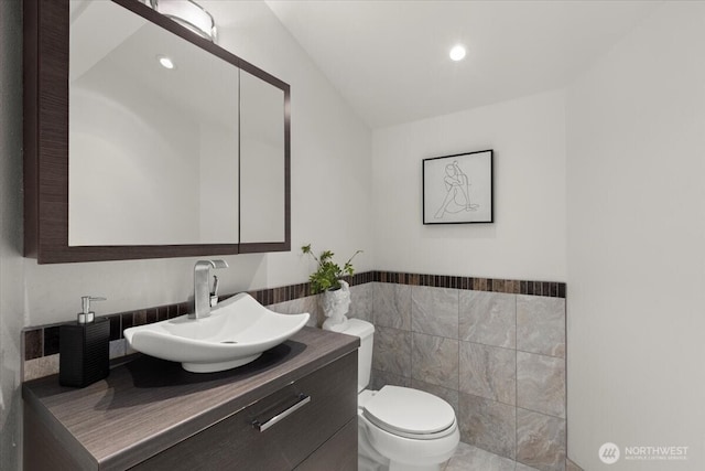 half bathroom with tile walls, toilet, vanity, a wainscoted wall, and recessed lighting