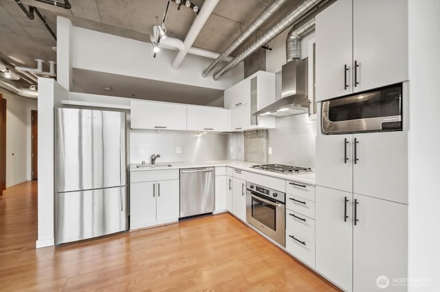 kitchen with stainless steel appliances, wall chimney exhaust hood, a sink, light wood-style flooring, and a high ceiling