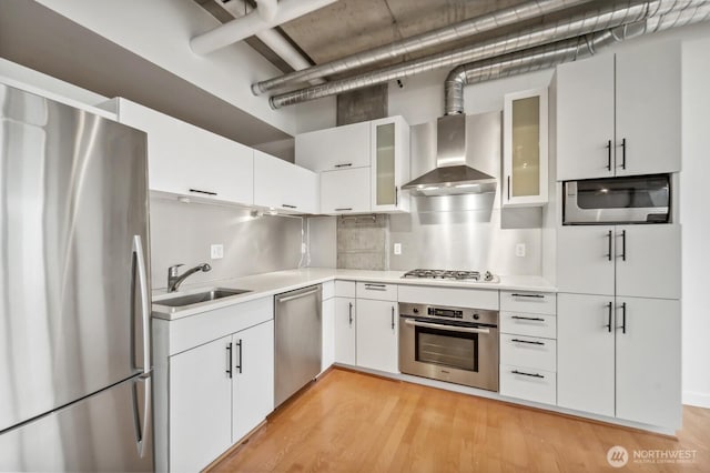 kitchen featuring appliances with stainless steel finishes, a sink, wall chimney exhaust hood, light countertops, and light wood-style flooring