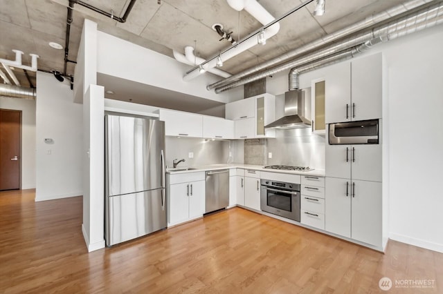 kitchen featuring stainless steel appliances, wall chimney range hood, a sink, light countertops, and white cabinets