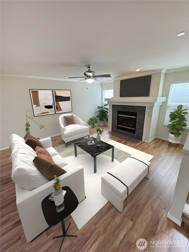 living area featuring crown molding, wood finished floors, a ceiling fan, a tiled fireplace, and baseboards