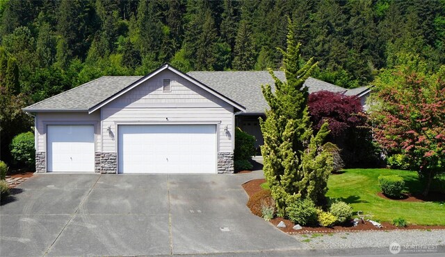 single story home featuring roof with shingles, stone siding, a garage, and a forest view