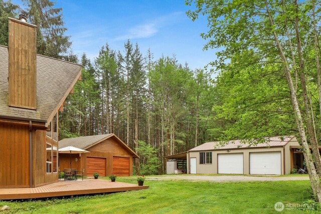 view of grassy yard featuring a detached garage, a wooden deck, an outbuilding, and a view of trees