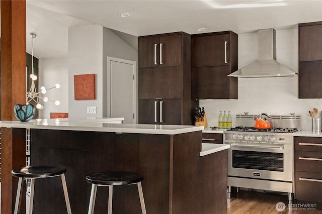 kitchen with wall chimney range hood, high end stainless steel range oven, a kitchen breakfast bar, light countertops, and dark brown cabinetry