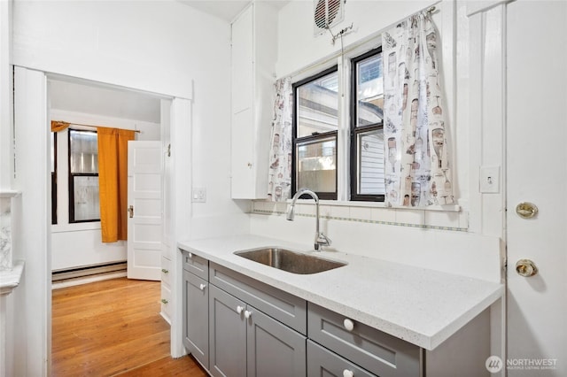 kitchen featuring a sink, gray cabinets, baseboard heating, light wood-style floors, and light stone countertops