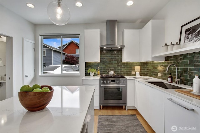kitchen with wall chimney exhaust hood, stainless steel range, a sink, white cabinets, and backsplash