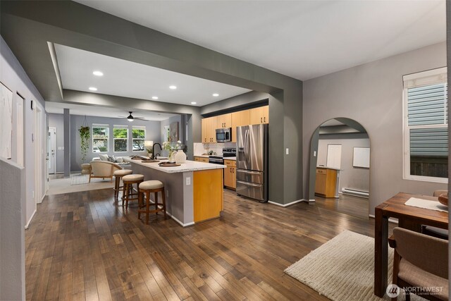 kitchen with stainless steel appliances, open floor plan, a kitchen island with sink, arched walkways, and dark wood-type flooring