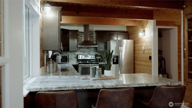 kitchen with stainless steel appliances, wall chimney range hood, a peninsula, light stone counters, and a kitchen breakfast bar