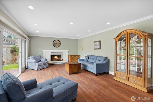 living room featuring wood finished floors, recessed lighting, ornamental molding, a warm lit fireplace, and baseboards