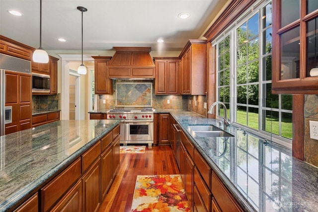 kitchen with built in appliances, a sink, premium range hood, dark wood finished floors, and decorative light fixtures