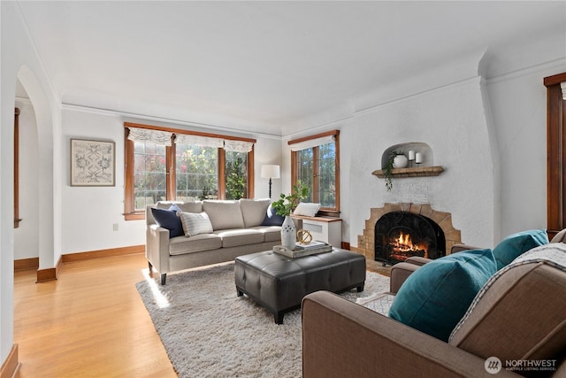 living area with a warm lit fireplace, arched walkways, light wood-style flooring, ornamental molding, and baseboards