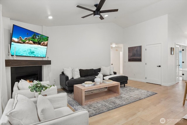 living area with ceiling fan, light wood-type flooring, vaulted ceiling, a lit fireplace, and baseboards
