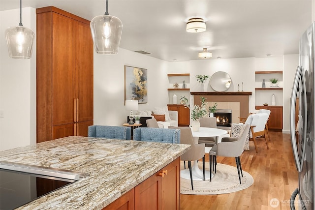 kitchen with freestanding refrigerator, light wood-style flooring, a fireplace, brown cabinetry, and hanging light fixtures