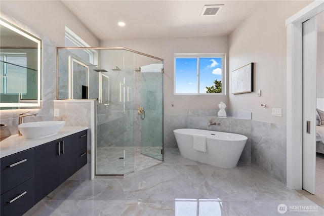 ensuite bathroom featuring tile walls, marble tiled flooring, a stall shower, a soaking tub, and vanity