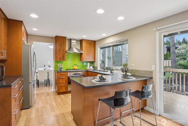kitchen with stainless steel appliances, wall chimney range hood, brown cabinetry, a sink, and a peninsula