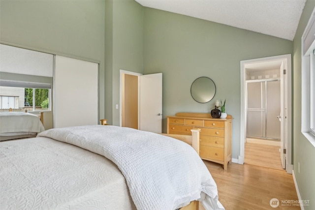 bedroom featuring light wood finished floors, a closet, high vaulted ceiling, baseboards, and ensuite bath