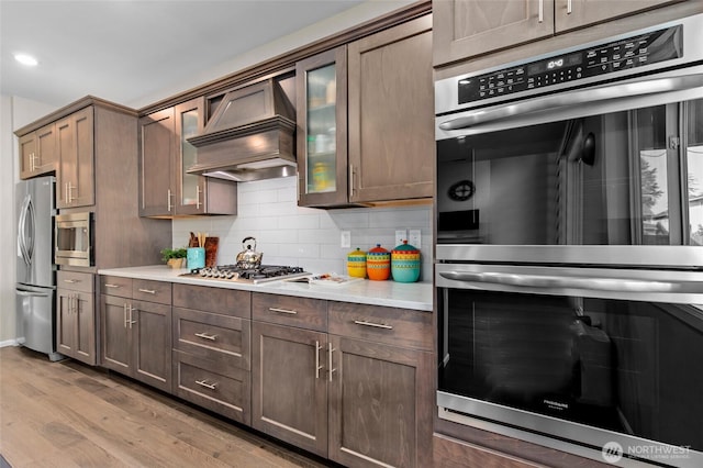 kitchen featuring stainless steel appliances, premium range hood, wood finished floors, light countertops, and decorative backsplash