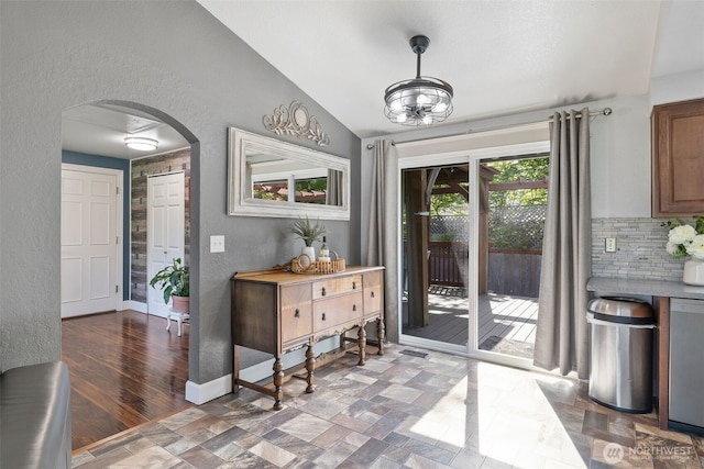 doorway to outside with arched walkways, a textured wall, vaulted ceiling, baseboards, and stone finish flooring