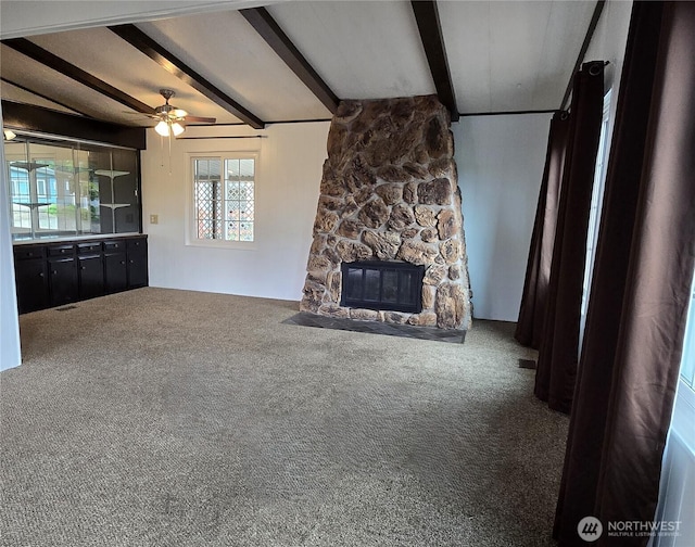 unfurnished living room with beamed ceiling, ceiling fan, carpet floors, and a stone fireplace