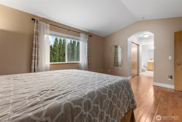 bedroom with light wood-style flooring, vaulted ceiling, arched walkways, baseboards, and ensuite bath
