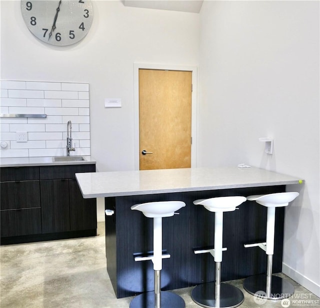 kitchen with a sink, a breakfast bar area, backsplash, light countertops, and concrete floors