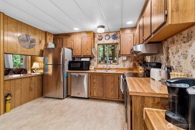 kitchen with black appliances, a sink, under cabinet range hood, wood counters, and brown cabinetry