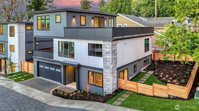 modern home with an attached garage, fence, decorative driveway, a balcony, and stone siding