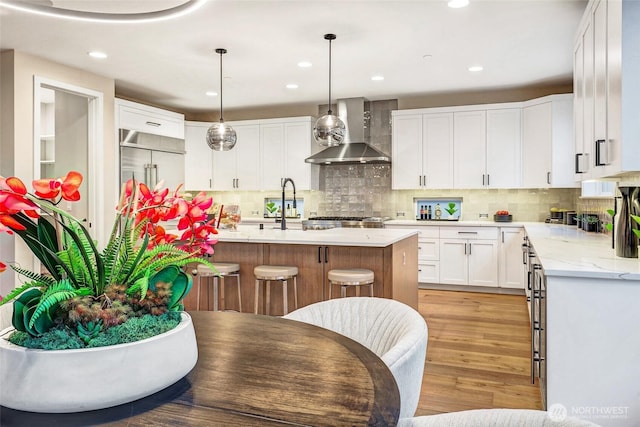 kitchen with wall chimney range hood, a kitchen island with sink, a sink, decorative backsplash, and white cabinetry