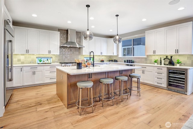 kitchen featuring beverage cooler, wall chimney range hood, light wood-style floors, a center island with sink, and white cabinetry