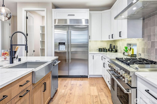 kitchen with high quality appliances, wall chimney range hood, a sink, light wood-style flooring, and backsplash