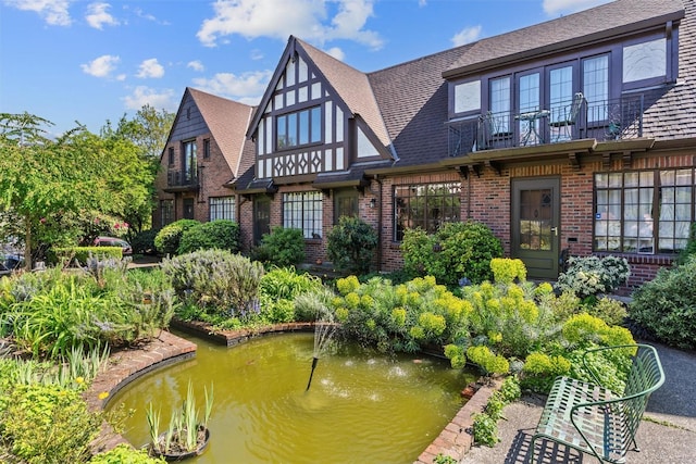 back of property with brick siding, a balcony, a small pond, and a shingled roof