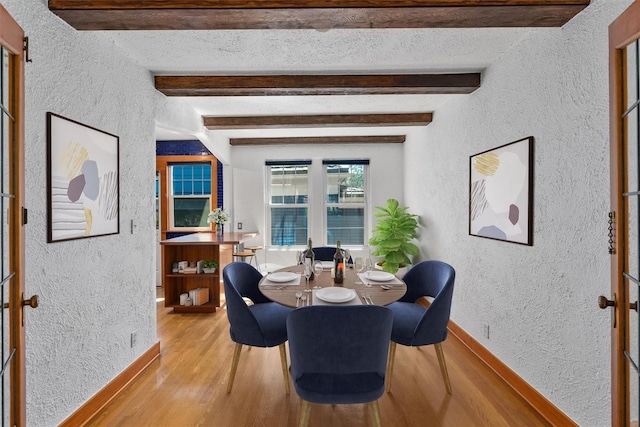 dining area featuring a textured wall, light wood finished floors, beamed ceiling, and baseboards