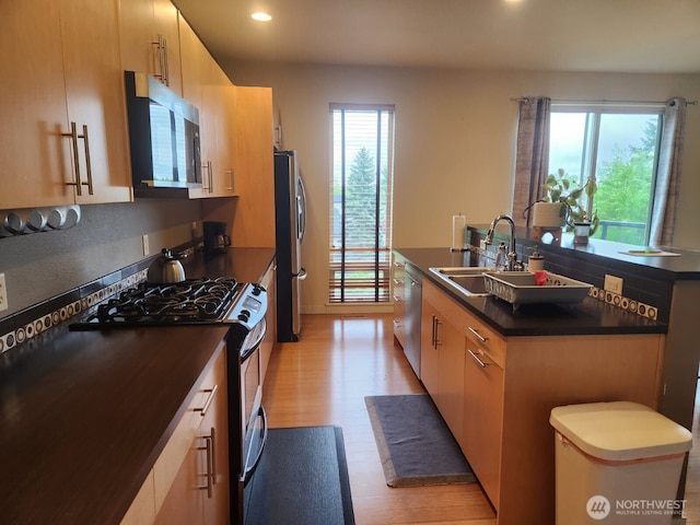 kitchen featuring appliances with stainless steel finishes, a sink, an island with sink, dark countertops, and light wood-style floors