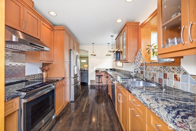 kitchen with under cabinet range hood, appliances with stainless steel finishes, a sink, brown cabinetry, and dark stone counters