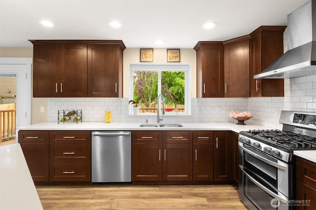 kitchen featuring stainless steel appliances, wall chimney exhaust hood, a sink, light countertops, and recessed lighting