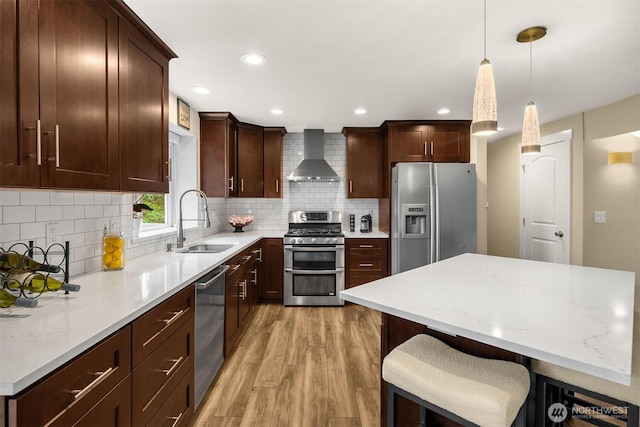 kitchen with appliances with stainless steel finishes, wall chimney range hood, light wood-type flooring, a sink, and pendant lighting