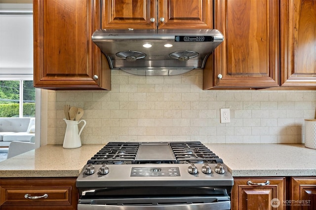 kitchen with stainless steel range with gas cooktop, under cabinet range hood, decorative backsplash, brown cabinetry, and light stone countertops