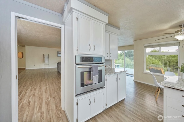 kitchen featuring stainless steel oven, a textured ceiling, a ceiling fan, light wood-style floors, and white cabinetry