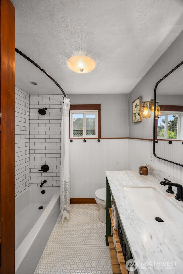 full bath featuring toilet, plenty of natural light, shower / bath combination with curtain, vanity, and a wainscoted wall