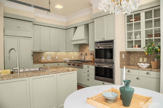 kitchen with appliances with stainless steel finishes, custom range hood, a chandelier, a sink, and glass insert cabinets